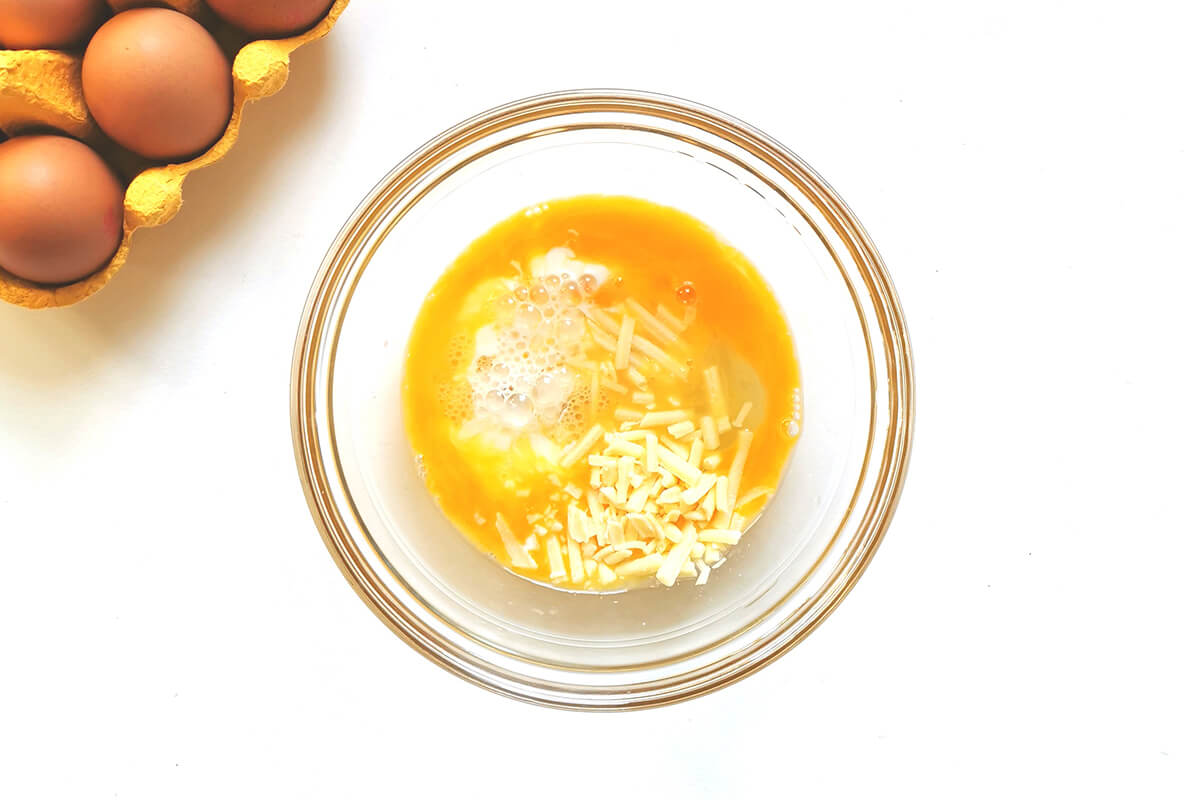 A glass bowl of whisked eggs with milk and cheese, next to a crate of eggs