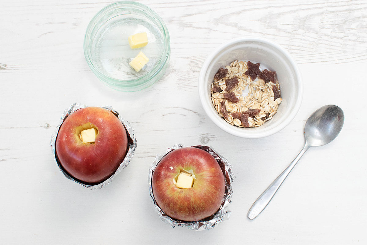 2 cored apples with foil wrapped around the bottom half of them, filled with oats and Organix Goodies, with a small square of butter on top