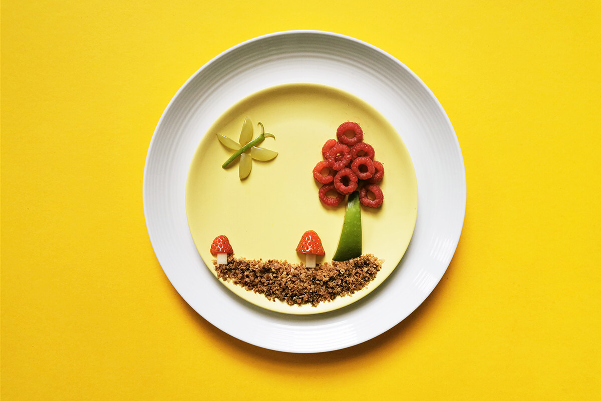 Gruffalo Biscuit Fun Plate, decorated with crumbled biscuits, an apple slice and raspberries to make a tree, apple chunks and strawberries to make 2 toadstools and an apple slice and grapes pieces to make a butterfly