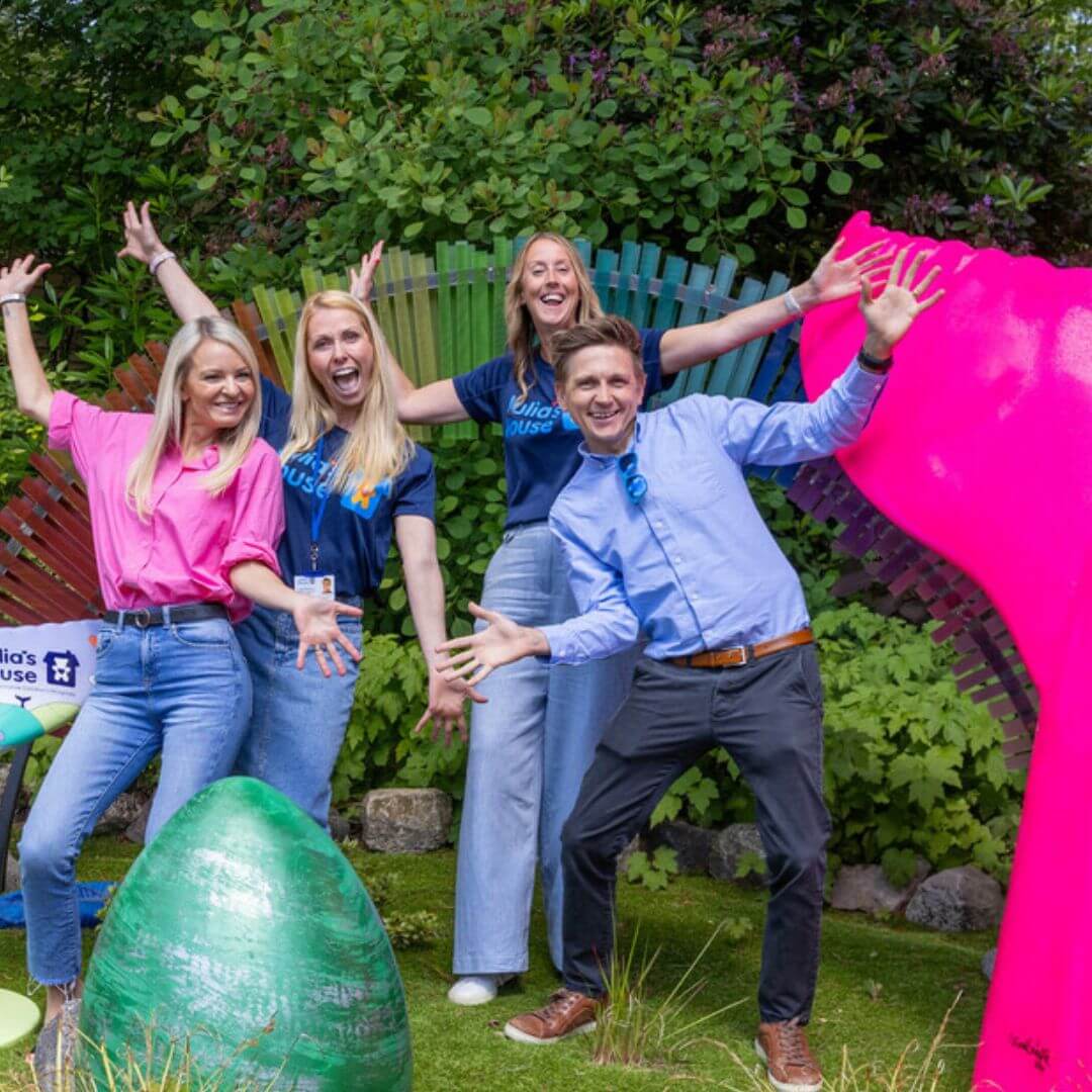 4 people smiling next to two sculpture, a pink mermaid tail and a large egg. They are outside amongst greenery.