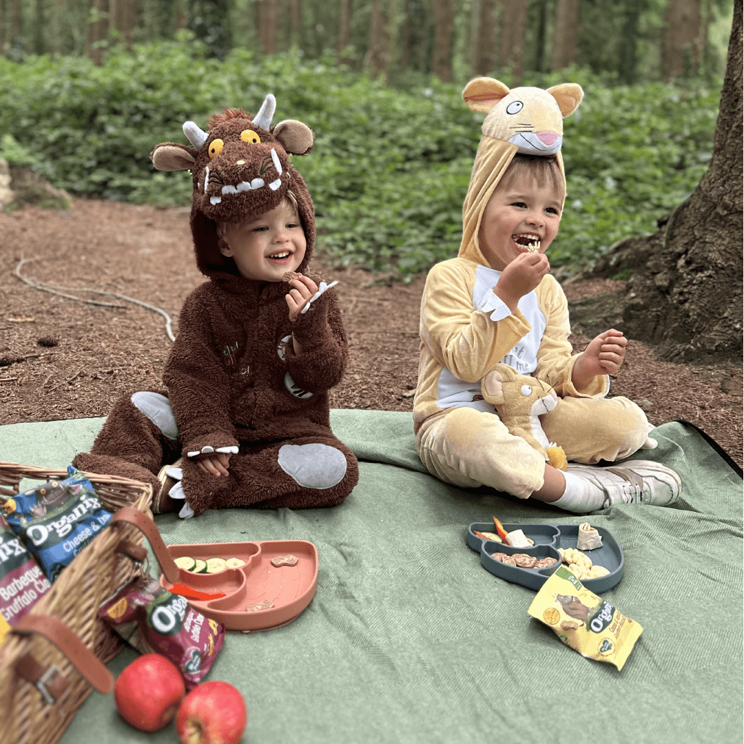 Two toddlers dressed in Gruffalo and mouse costumes sit on a picnic blanket in the woods, smiling and snacking on Organix treats from lunchboxes and a picnic basket.