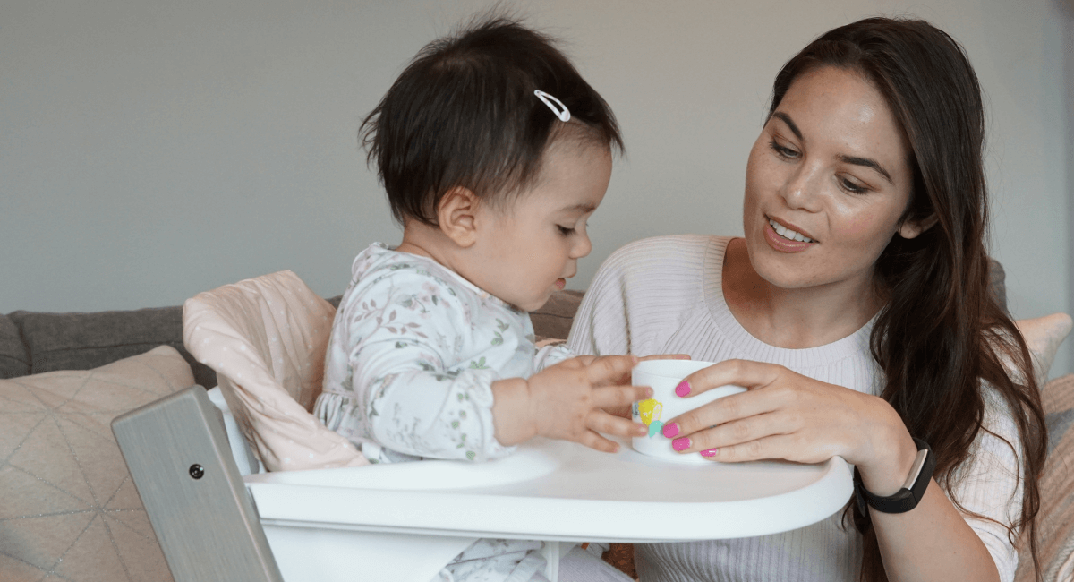 Mother crouched down with baby daughter in a high chair. Both looking and smiling at each other whilst holding a cup.