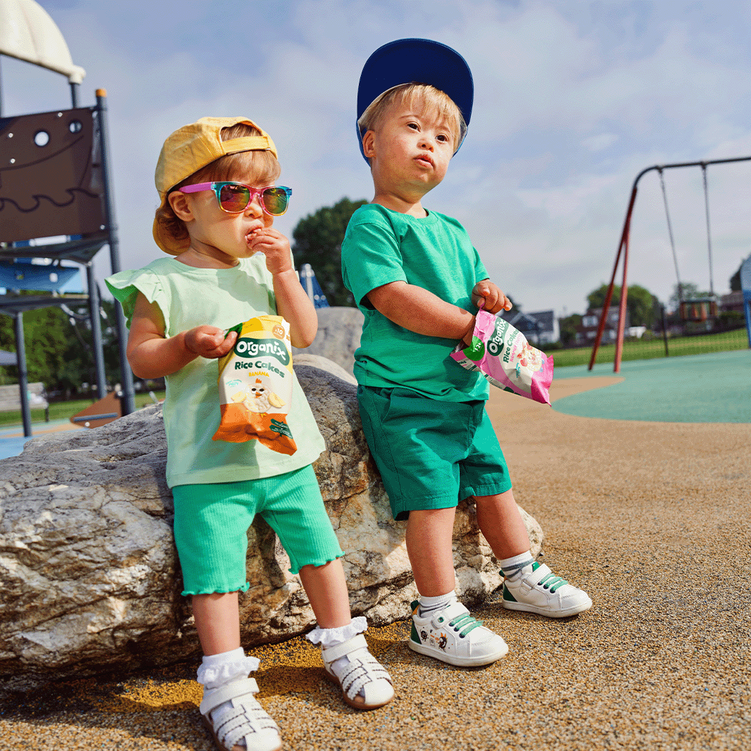 Two young children standing and eating Organix snacks. They are holding the snacks in their hands, with the Organix branding visible. The setting is bright and casual, emphasizing healthy, organic snacking and an active lifestyle.