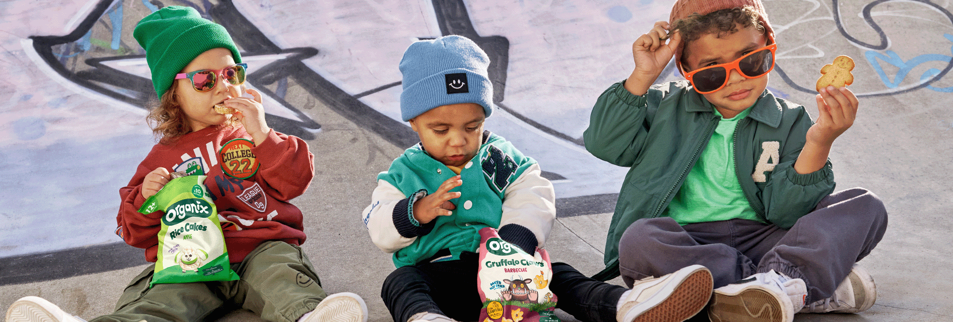 3 toddlers in a skate park in cool clothing and beanie hats each eating different Organix snacks. Apple Rice Cakes, BBQ Claws and Gingerbread men biscuits.
