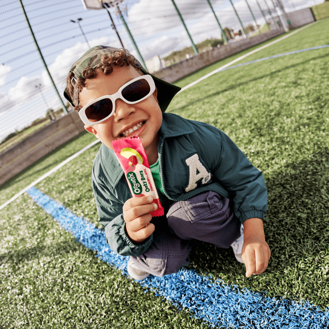 Toddler Boy holding an Organix Strawberry and Apple Oaty Bar in a grass outdoor basketball court. He is wearing a backwards cap and sunglasses with a big smile