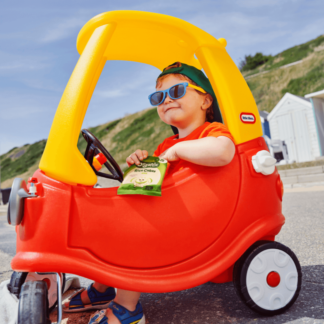 Toddler boy eating Organix apple rice cake clouds on the beach in a red and yellow plastic car, wearing a backwards cap and sunglasses