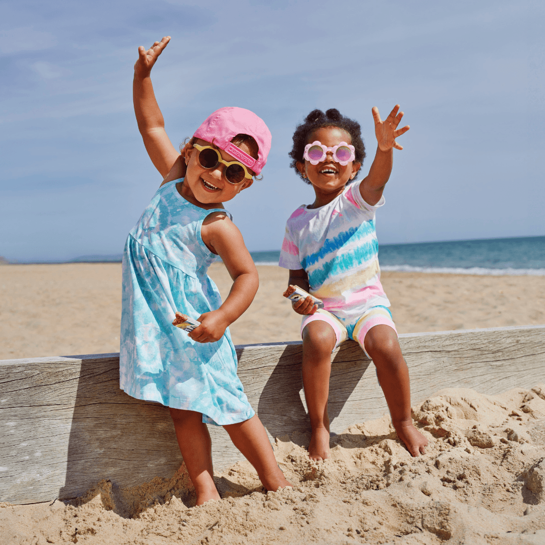 Two girls, toddlers, eating Organix Oaty bars in the beach in summer dresses and funky sunglasses. Looking happy