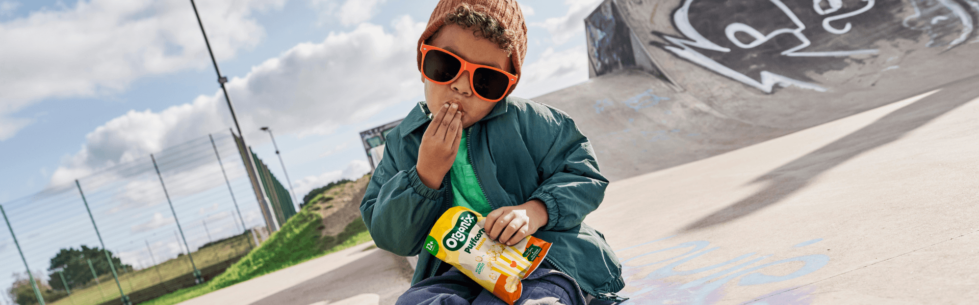 Toddler boy eating Organix Banana Puffcorn in a skatepark wearing cool sunglasses and an orange beanie.